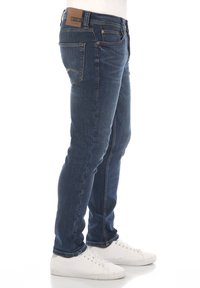 Jeans de mezclilla azul oscuro con un diseño ajustado, cinco bolsillos y una etiqueta de cuero en la parte trasera de la cinturilla. Combinados con zapatillas blancas.