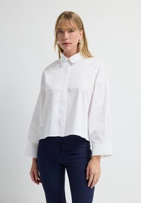 Camisa blanca corta con cuello de botón, mangas largas y detalle en la costura delantera. Combinada con pantalones ajustados azules marino.