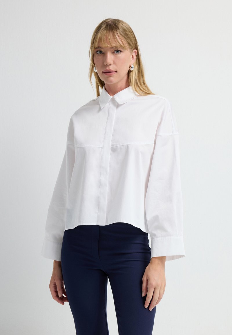 Camisa blanca corta con cuello de botón, mangas largas y detalle en la costura delantera. Combinada con pantalones ajustados azules marino.