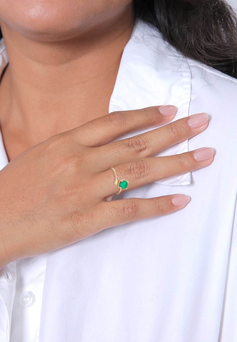 Anello d'oro con una pietra verde, caratterizzato da un design ondulato. Dito adornato, con unghie lucidate di colore chiaro, su uno sfondo di camicia bianca.