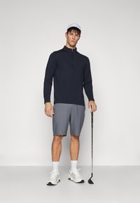 Marine langermet glidelås topp, grå shorts, hvite sneakers, hvit caps, holder en golfkølle, står mot en enkel lys bakgrunn.