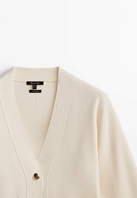 Krämfärgad cardigan med V-ringning, tillverkad av mjukt tyg. Har en enkel knappstängning och en subtil textur. Märket inuti synligt.