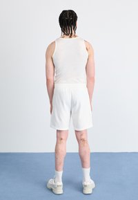 White tank top and shorts, with visible skin texture. Tan and brown patterns on legs. White sneakers and socks. Plain background.