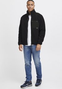 Zwarte fleece jacket met een hoge kraag, een ritsborstzak en een gestructureerd ontwerp, gecombineerd met blauwe jeans en donkere sneakers.