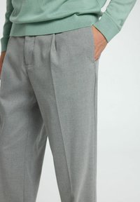Pantalons taillés gris clair avec poches latérales, présentant une texture de tissu lisse et des plis plats. Associés à un pull vert menthe.