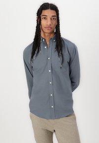 Camisa de botones gris con un bolsillo frontal, manga larga y un dobladillo curvado. La tela parece suave y ligera.