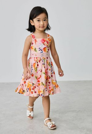 Junges Mädchen, das in einem rosa Kleid mit bunten Fruchtmotiven und weißen Sandalen mit Schleifen auf einem schlichten grauen Boden läuft.