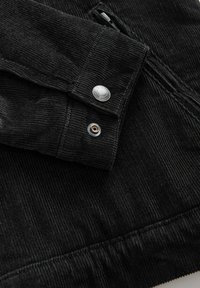 Zwarte corduroy jas-mouw met zilveren drukknoop en zichtbare stiksels op de gestructureerde stof.