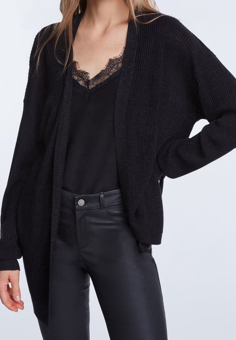 Femme portant un caraco noir bordé de dentelle, un cardigan noir côtelé et un pantalon en cuir noir taille haute, une main sur la hanche.