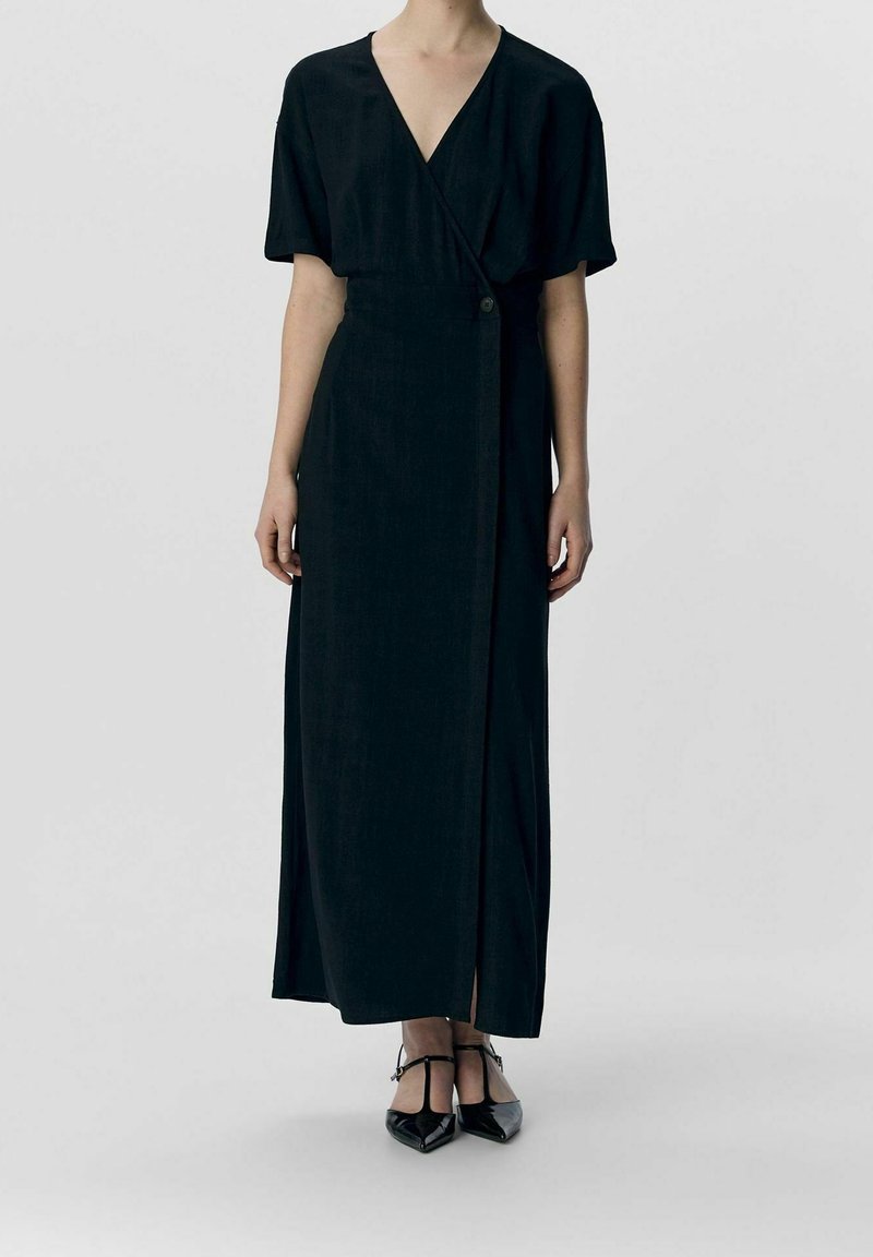 Femme portant une robe portefeuille noire à manches courtes et col en V, avec un bouton à la taille, associée à des talons noirs pointus à bride en T.