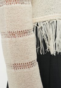 Ανοιχτό πλεκτό, εκρού σάλι με κρόσσια στα άκρα και οριζόντια ριγέ μοτίβα, με υφή από νήμα. Φορεμένο πάνω από σκούρο τοπ.