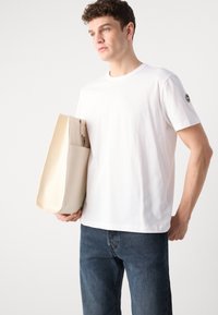White cotton t-shirt with short sleeves, round neckline, and a logo on the left sleeve. The model holds a beige canvas tote bag.