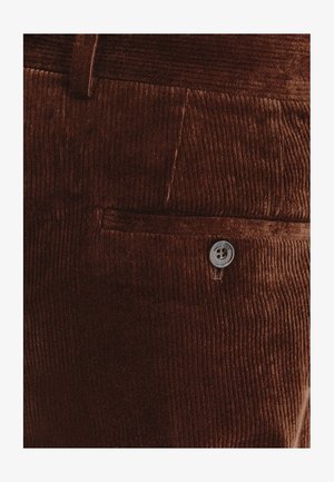 Brązowa tkanina sztruksowa z pionowym żeberkowaniem, z boczną kieszenią i zapięciem na srebrny guzik. Miękka tekstura i usztywniony design.