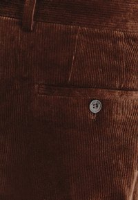 Brązowa tkanina sztruksowa z pionowym żeberkowaniem, z boczną kieszenią i zapięciem na srebrny guzik. Miękka tekstura i usztywniony design.