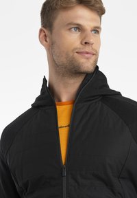 Black zip-up jacket with a hood, featuring textured fabric and solid color accents, worn over an orange shirt. Minimalist design.