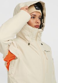 Lichtbeige waterproof jack met een hoge kraag, ritssluiting aan de voorkant en oranje mesh voering zichtbaar in de mouw. Heeft een verstelbare capuchon en merkname.