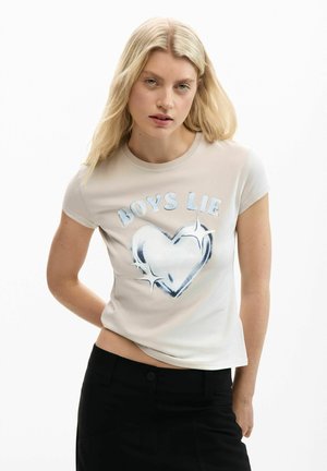 Blondínka v béžovom tričku s trblietavým srdcovým motívom a nápisom "CHLAPCI KLAMÚ", v čiernych nohaviciach, na bielom pozadí.