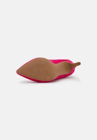 Chaussure à talon haut pointu en daim rose, couchée sur le côté, montrant une semelle lisse couleur fauve et un talon aiguille fin sur fond blanc.