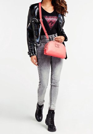 Femme portant une veste en cuir verni noir, un t-shirt graphique noir, un jean skinny gris délavé, des bottes de combat noires et portant un sac bandoulière corail.