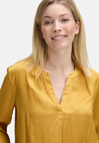Lächelnde Frau mit glattem blonden Haar, die eine senfgelbe Bluse trägt, vor einem einfarbigen Hintergrund.