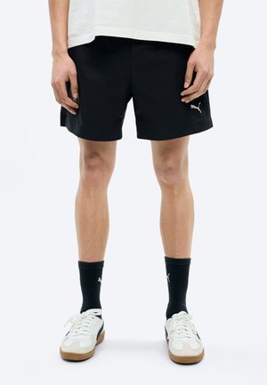 Osoba oblečená v čiernych šortkách Puma, čiernych ponožkách Puma a bielych teniskách Puma, stojaca pred jednoduchým bielym pozadím.