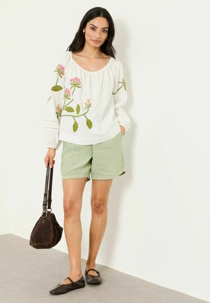 Žena v bielej blúzke s kvetinovou výšivkou, svetlozelených šortkách, čiernych balerínach a s tmavohnedou kabelkou po boku.