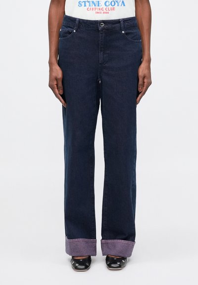 Jean large bleu foncé avec une fermeture à bouton, cinq poches et des revers de pantalon avec un accent violet. Fabriqué en tissu denim.