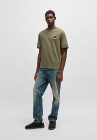 Oliven grøn bomulds t-shirt med korte ærmer og et gult grafisk design på brystet, parret med slidte blå jeans og sorte sneakers.