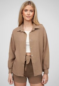 Camicia button-up marrone a trama con colletto, abbinata a un top corto bianco e pantaloni corti marroni coordinati con un dettaglio annodato in vita.