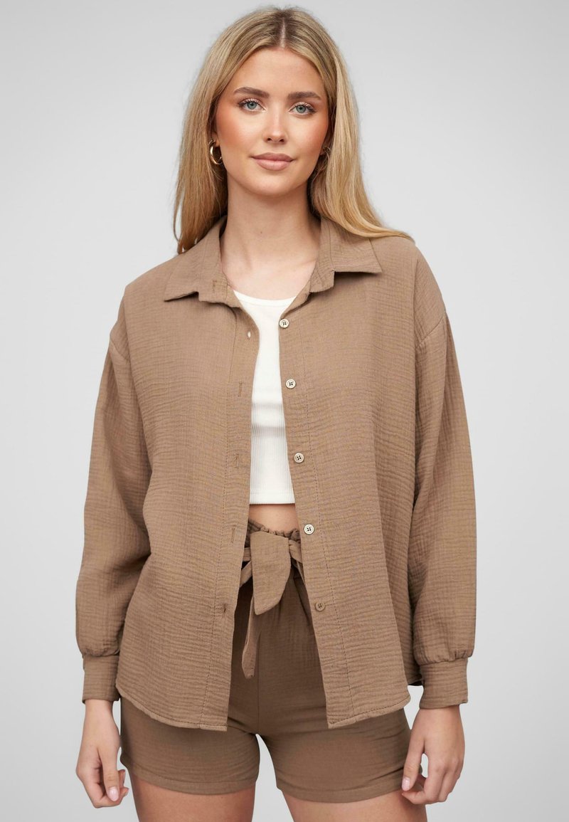 Camicia button-up marrone a trama con colletto, abbinata a un top corto bianco e pantaloni corti marroni coordinati con un dettaglio annodato in vita.