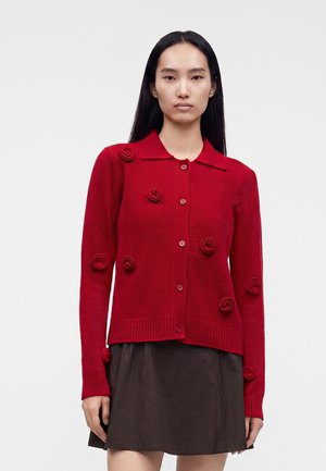 Femme portant un cardigan rouge boutonné avec des détails floraux texturés, associé à une jupe marron foncé, debout devant un fond uni.