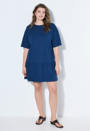 Młoda kobieta z falującymi włosami, ubrana w luźną granatową sukienkę z krótkim rękawem i czarne sandały, stojąca na białym tle.