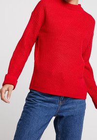 Röd stickad tröja med ribbat mönster, med långa ärmar och hög krage. I kombination med blå jeans.