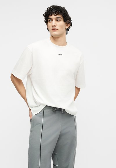 Νεαρός με σγουρά σκουρόχρωμα μαλλιά, φοράει μια λευκή φαρδιά μπλούζα με το κείμενο "OFF" και γκρι παντελόνι με μαύρες ραφές, κοιτάζει προς τα πλάγια.