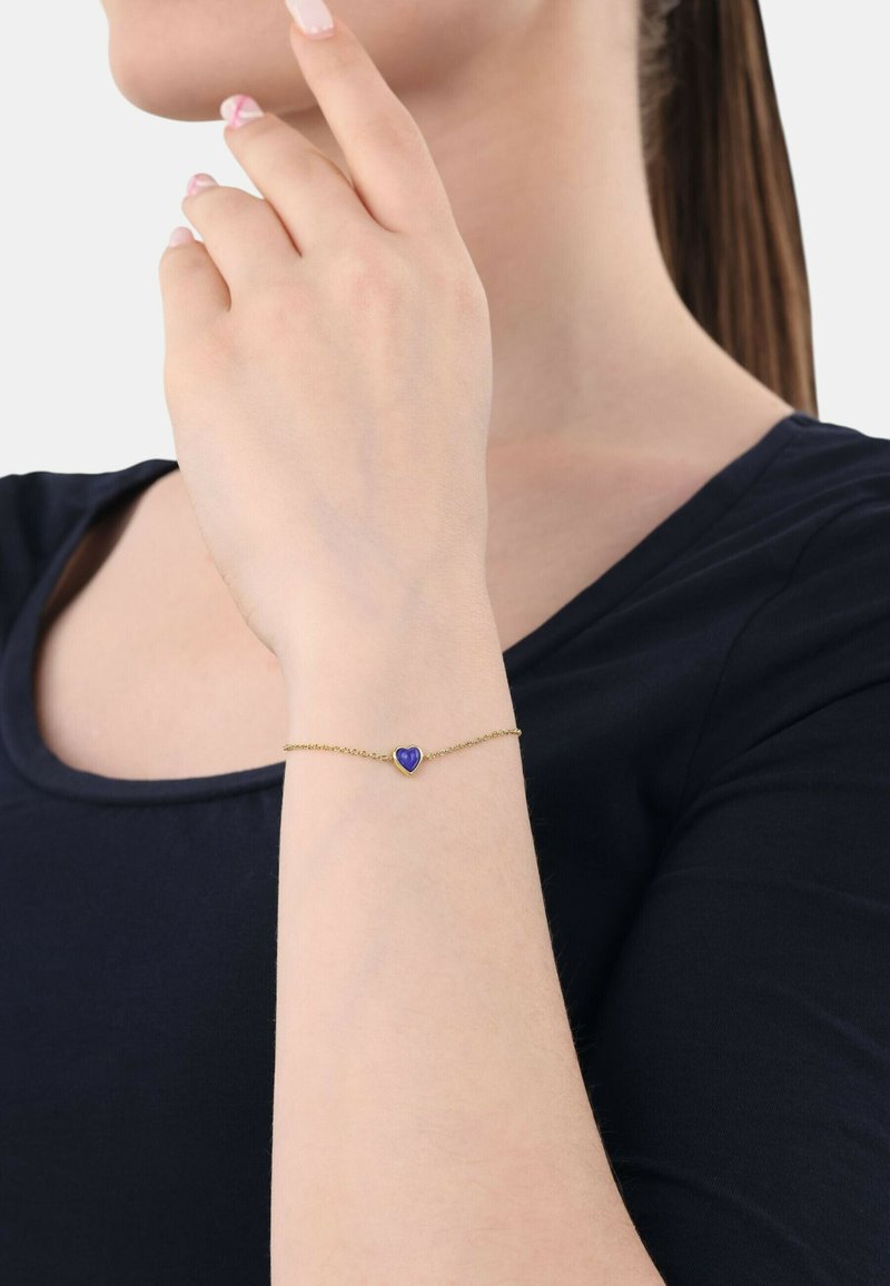 Bracciale in oro con un piccolo charm a forma di cuore blu, indossato su una mano, che evidenzia il design delicato del bracciale e il contrasto di colori.