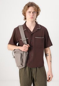 Kurzarmige braune Hemd mit weißem Piping, zwei Taschen und einem lässigen Schnitt. Graue Umhängetasche mit Reißverschluss und verstellbarem Tragegurt.