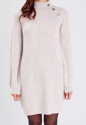 Woman wearing a light beige ribbed knit sweater dress with long sleeves and three decorative buttons near the shoulder.