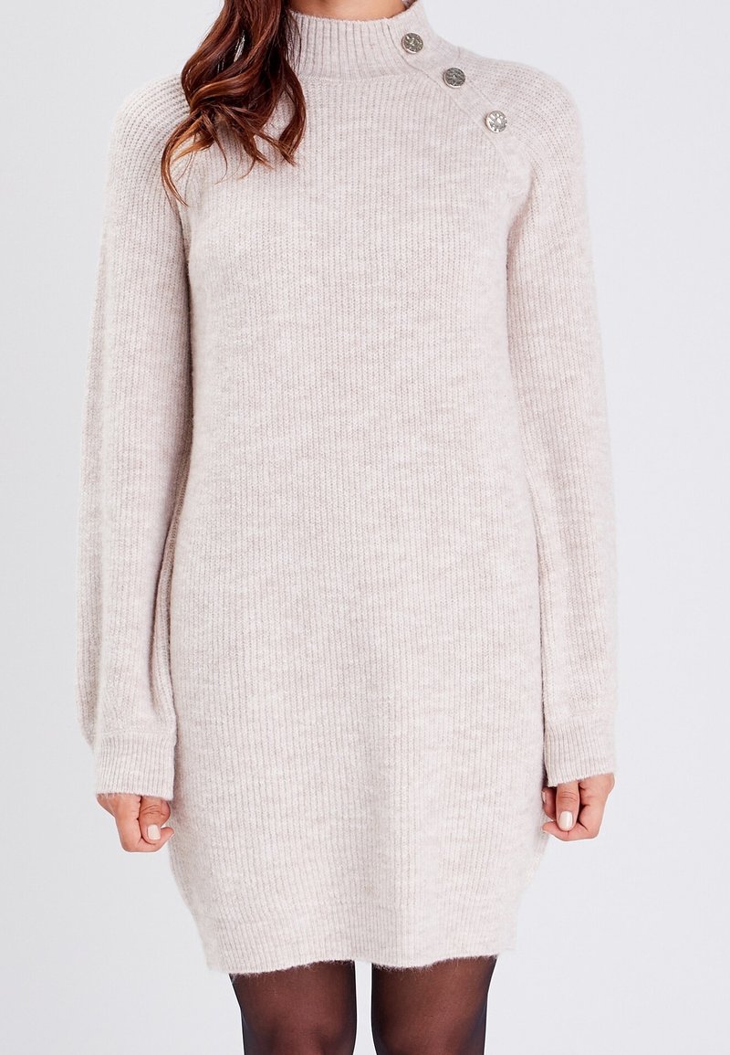 Femme portant une robe-pull côtelée beige clair à manches longues avec trois boutons décoratifs près de l'épaule.