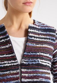 Chaqueta a rayas multicolor, con un tejido texturizado y bordes deshilachados, con cierre frontal de cremallera y cuello redondo. Llevada sobre una camiseta blanca.