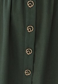 Tamsiai žalia, minkšta tekstūra pasižyminti medžiaga su daugybe auksinių apvalių sagų priekyje, suteikiančių kontrastinį detalę.