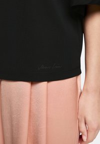 Black short-sleeve top with a smooth texture featuring a subtle logo near the hem, paired with a light pink pleated skirt.