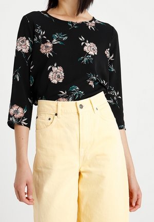 Blouse noire avec imprimé floral rose et manches trois-quarts, associée à un pantalon taille haute large, jaune pâle.