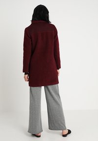 Burgunderfarbener, strukturierten Mantel mit einem Kragen, langen Ärmeln und gerader Silhouette, kombiniert mit grauen gemusterten, weiten Hosen.