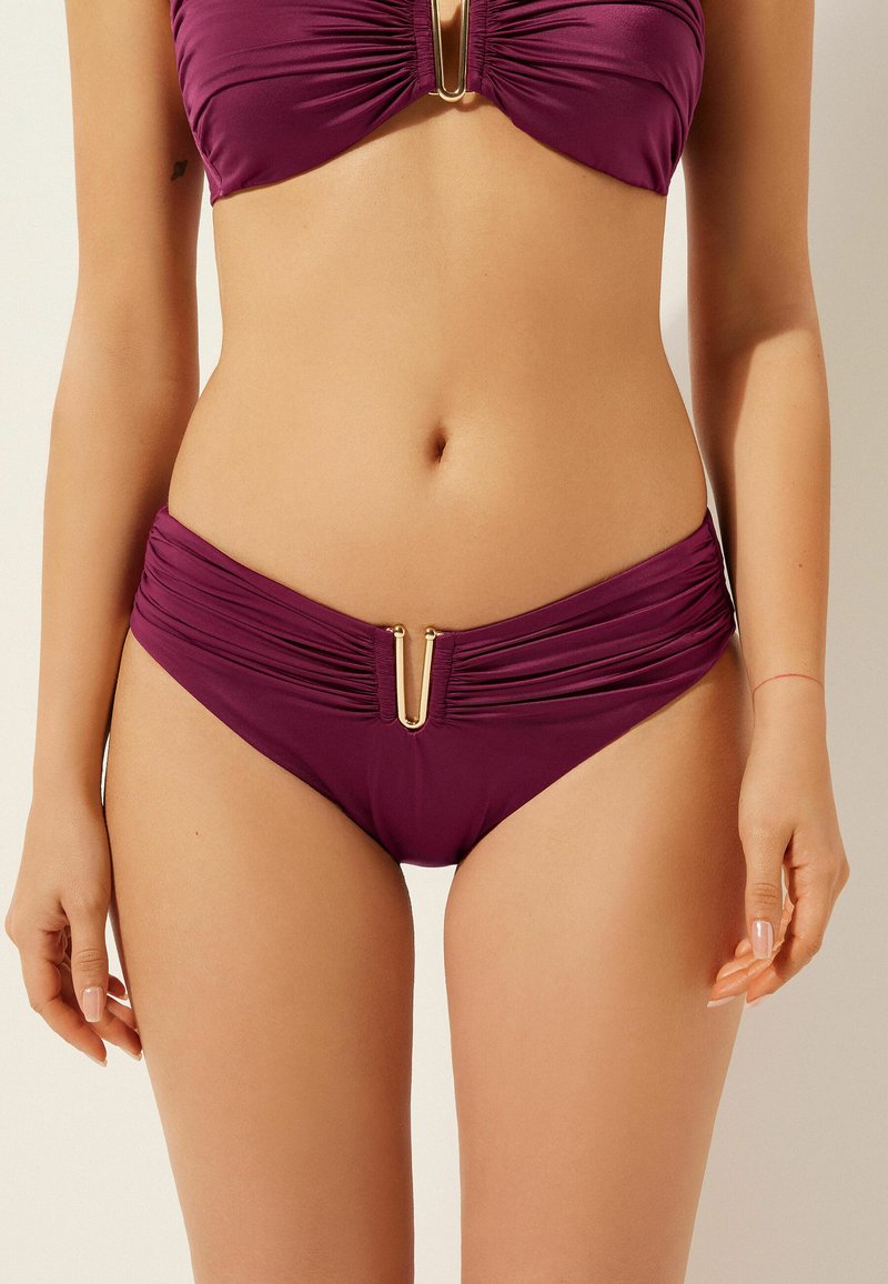 Bikini-set i djup lila tyg, med ruche-detaljer på både toppen och botten. Guldhardware pryder mitten av bh:n och midjebandet.