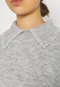 Gros plan sur un pull gris en maille avec un col pointu orné de deux rangées de petites perles blanches de chaque côté.