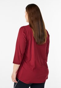 Bordeauxrotes Langarmshirt mit lockerem Schnitt und rundem Ausschnitt. Glatte Stoffstruktur und seitliche Einsätze mit geschwungenem Saum.