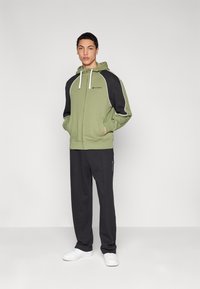 Sweat à capuche vert avec accents noirs, galon blanc et poches avant ; associé à un pantalon foncé et des baskets blanches. Design simple et sportif.