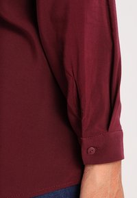 Primo piano di un braccio di una persona che indossa una camicia a maniche lunghe color bordeaux con il polsino abbottonato sopra dei jeans blu scuro.