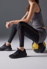 Mustat legginssit verkkopaneeleilla ja harmaa hihaton paita, mustien urheilukenkien kanssa, istuen keltaisella ja mustalla lääkepallolla.