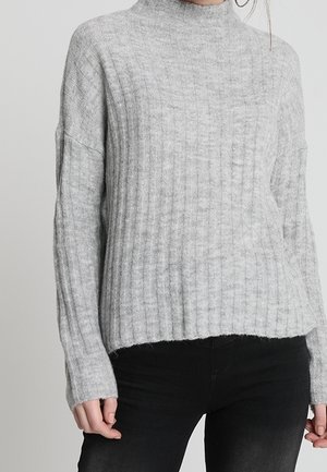 Femme portant un pull en maille côtelée gris clair à manches longues et un pantalon noir, debout devant un fond uni.